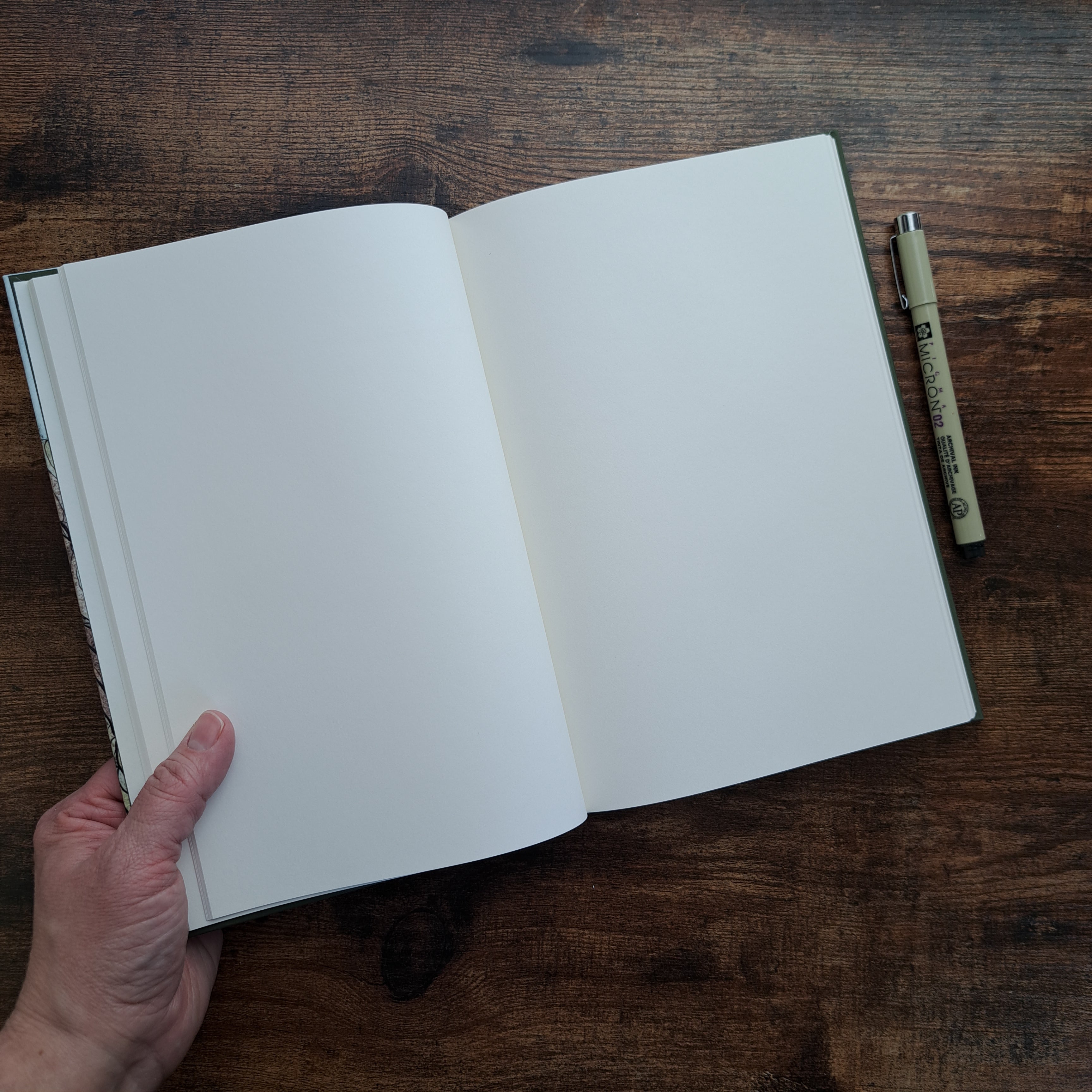 Open hardcover journal showing blank white pages, held by hand on wood surface with Micron pen


