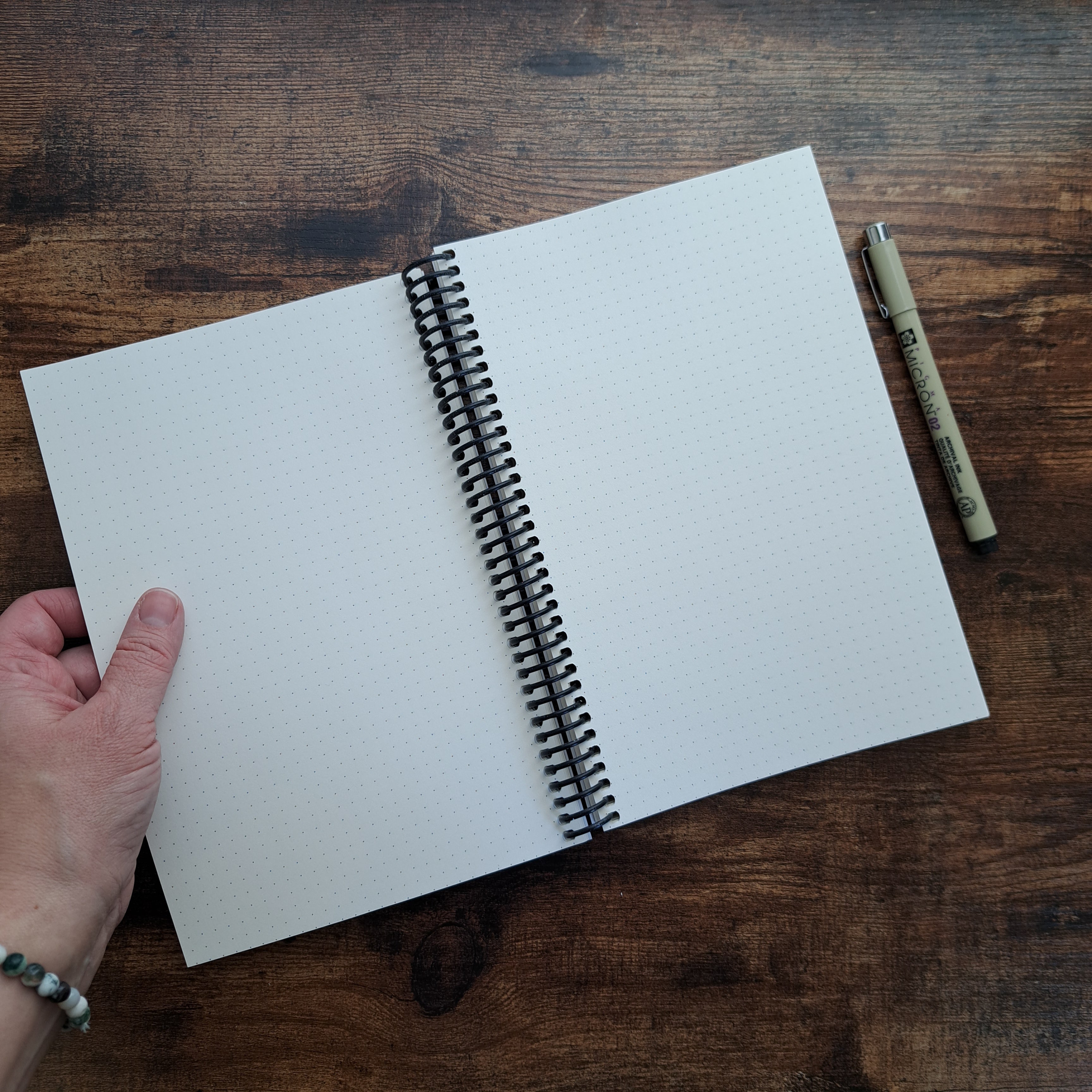 Open spiral-bound journal showing dot grid pages, held by hand on wood surface with Micron pen

